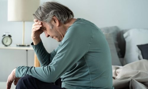 Old man holding his head with his hands sits on the bed
